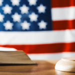 A gavel rests on a wooden block beside an open book in front of a blurred American flag backdrop. Accessibility icon is present.