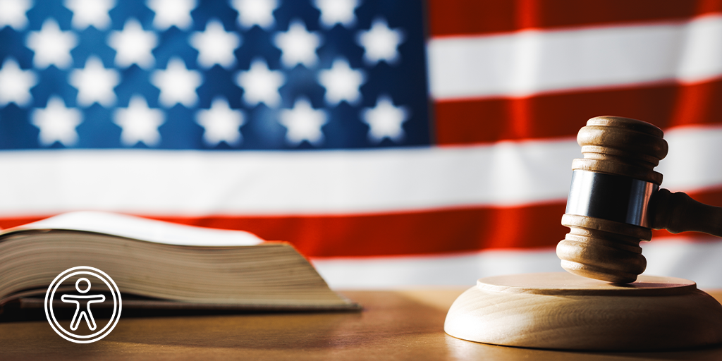 A gavel rests on a wooden block beside an open book in front of a blurred American flag backdrop. Accessibility icon is present.