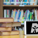 Stack of old hardcover books on a wooden table in front of a bookshelf filled with colorful books. An electronic tablet placed next to the books displays documenta11y logo on the screen.