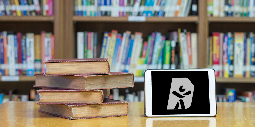 Stack of old hardcover books on a wooden table in front of a bookshelf filled with colorful books. An electronic tablet placed next to the books displays documenta11y logo on the screen.